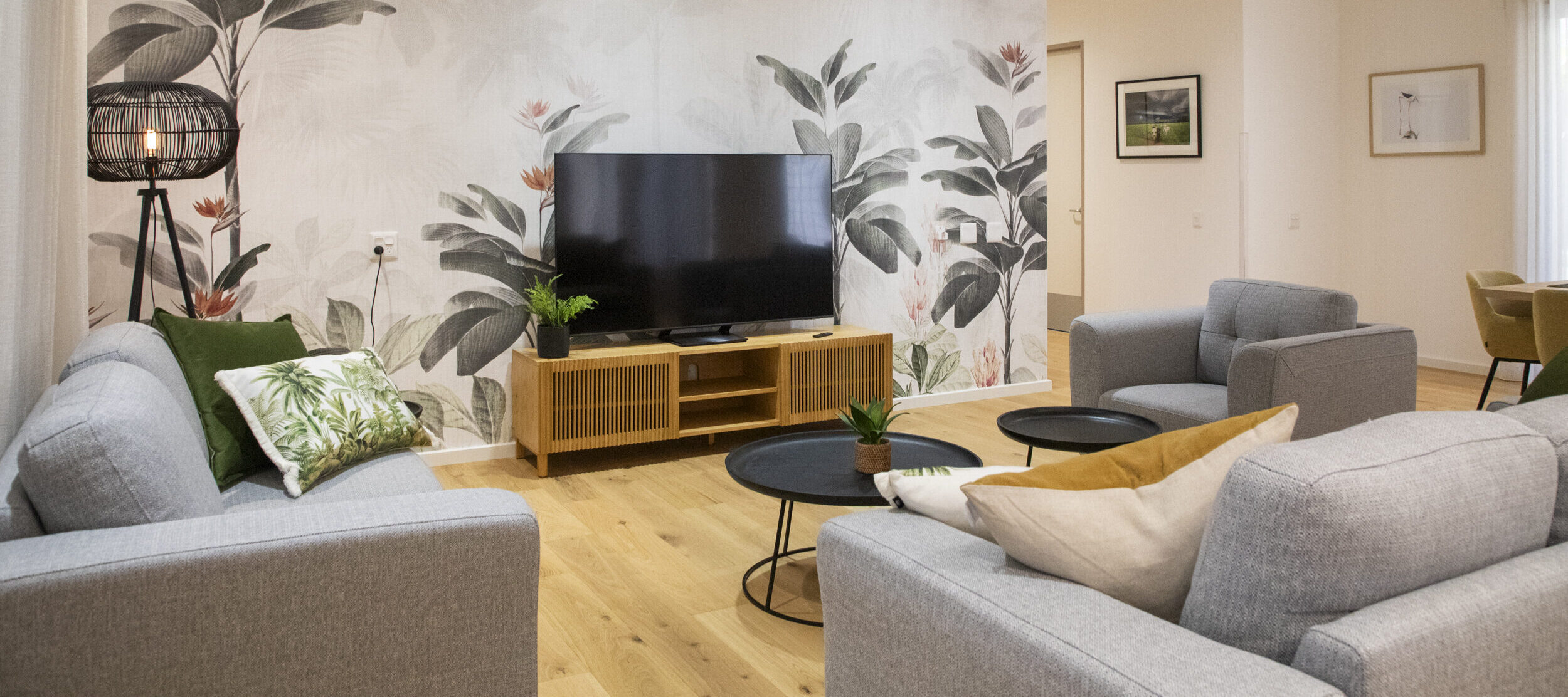 A wide angle of the lounge room of a Specialist Disability Accommodation home. Grey couches face a large black television which is in front of a patterned feature wall.