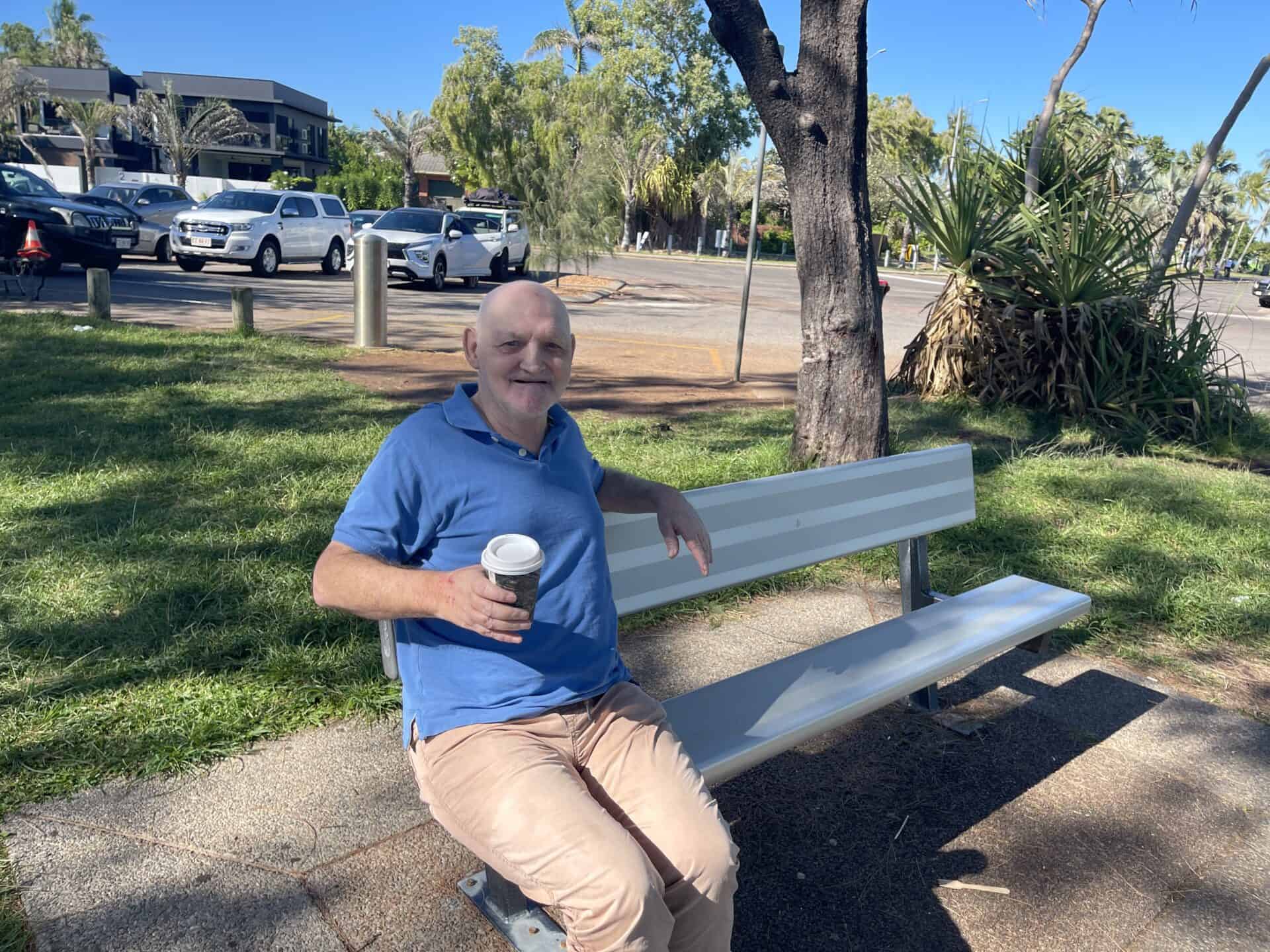 A man sits at one end of a long grey bench seat, holding a takeaway coffee cup. Several cars and trees are in the background.