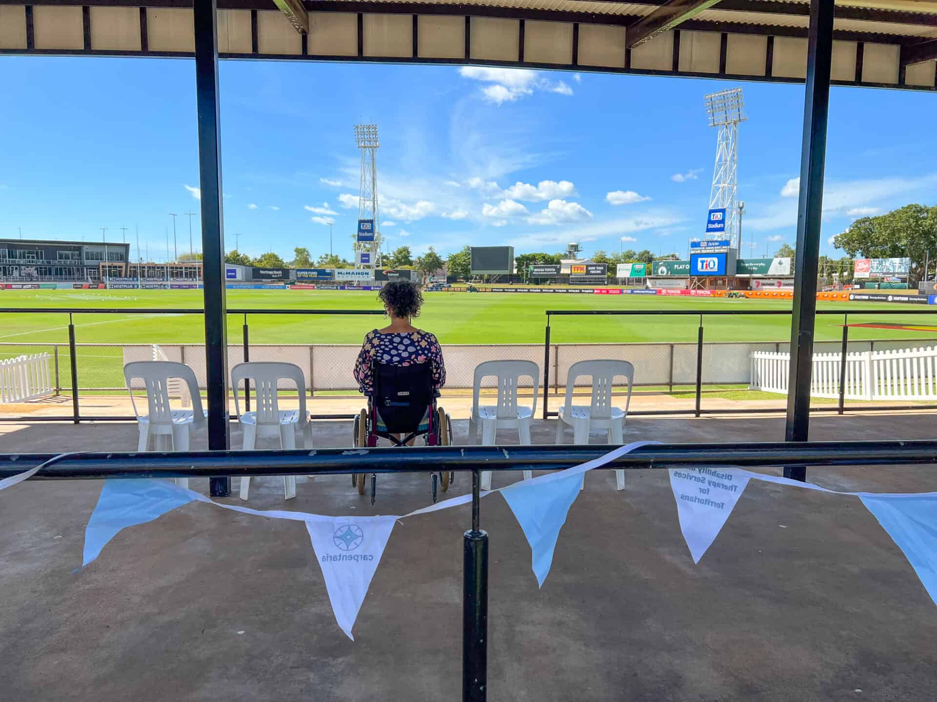 The TIO accessible viewing platform, feature a person sitting in a wheelchair looking out to the oval. Carpentaria bunting is on the rail in the foreground.