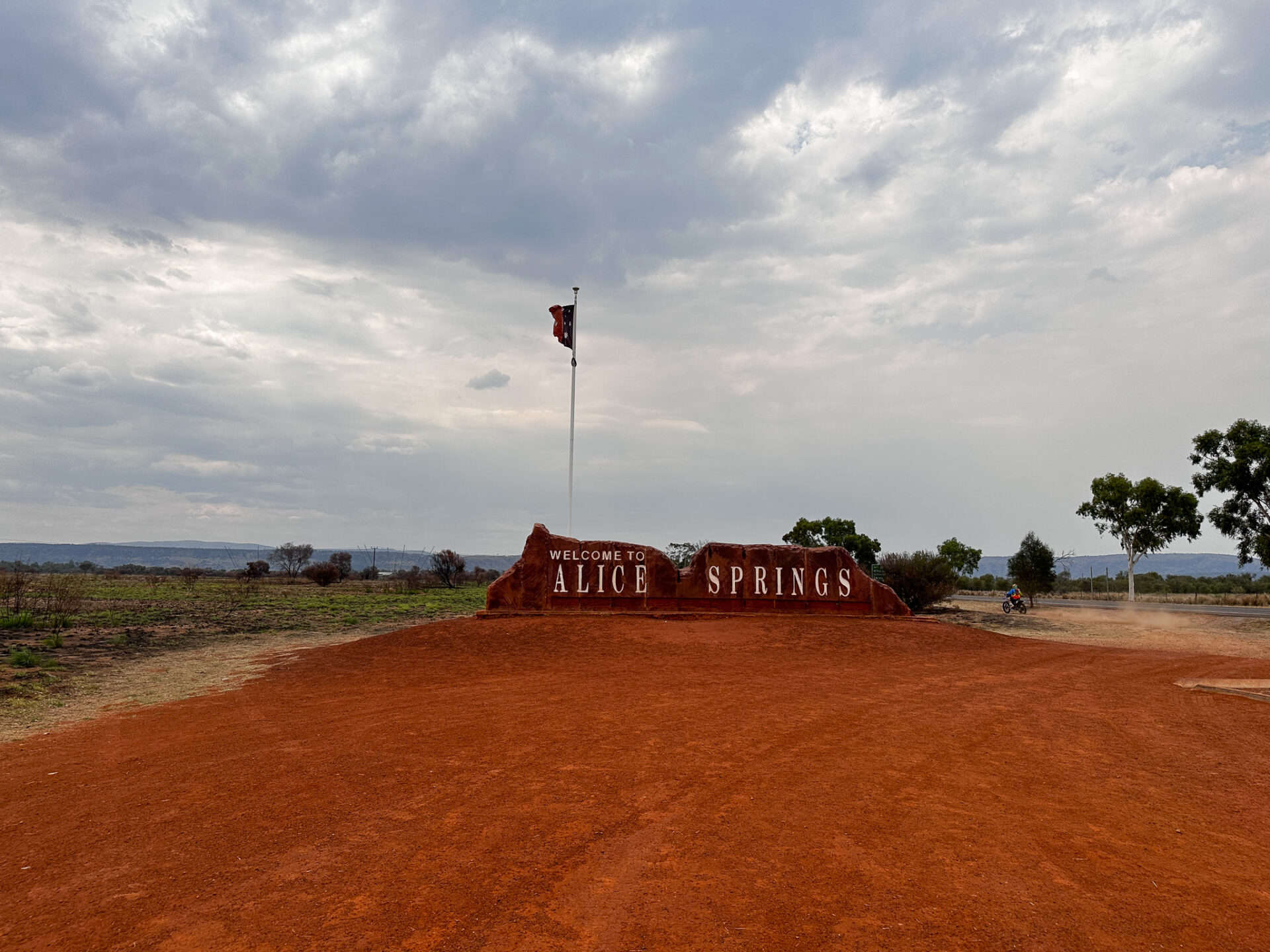 Text: 'Welcome to Alice Springs' is printed on a sign, in front of a flag pole. Red dirt fills the area in front of the sign, and some trees and grey sky is behind the sign.