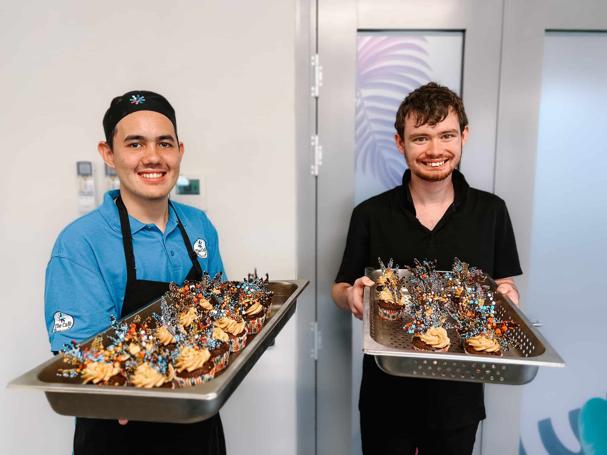 Two Cafe trainees hold trays of decorated cupcakes in the Cafe at Harry's Place.