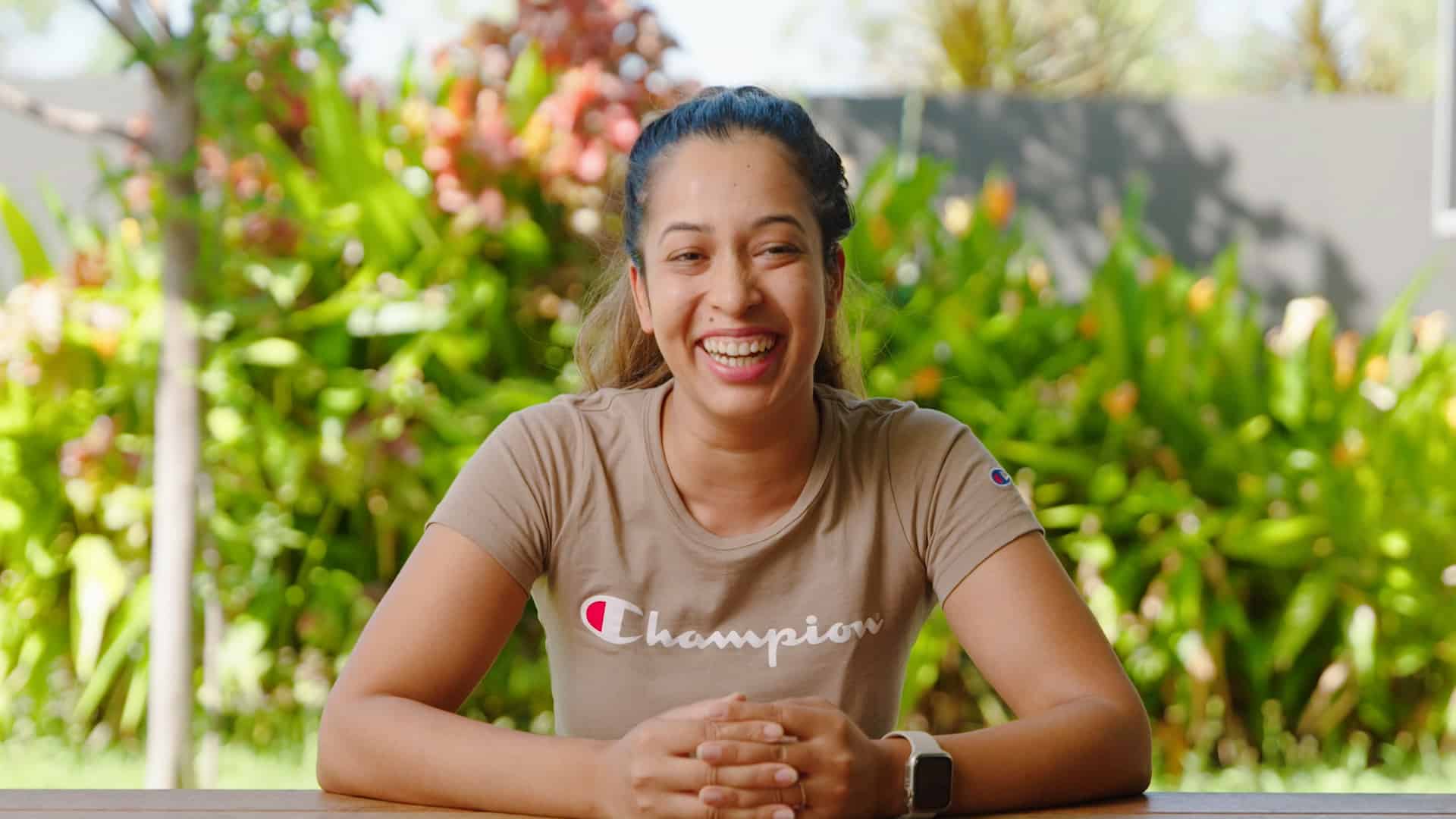 A young woman wearing a light brown t-shirt sits with her elbows on the table and smiles. Green shrubs are in the garden behind her.
