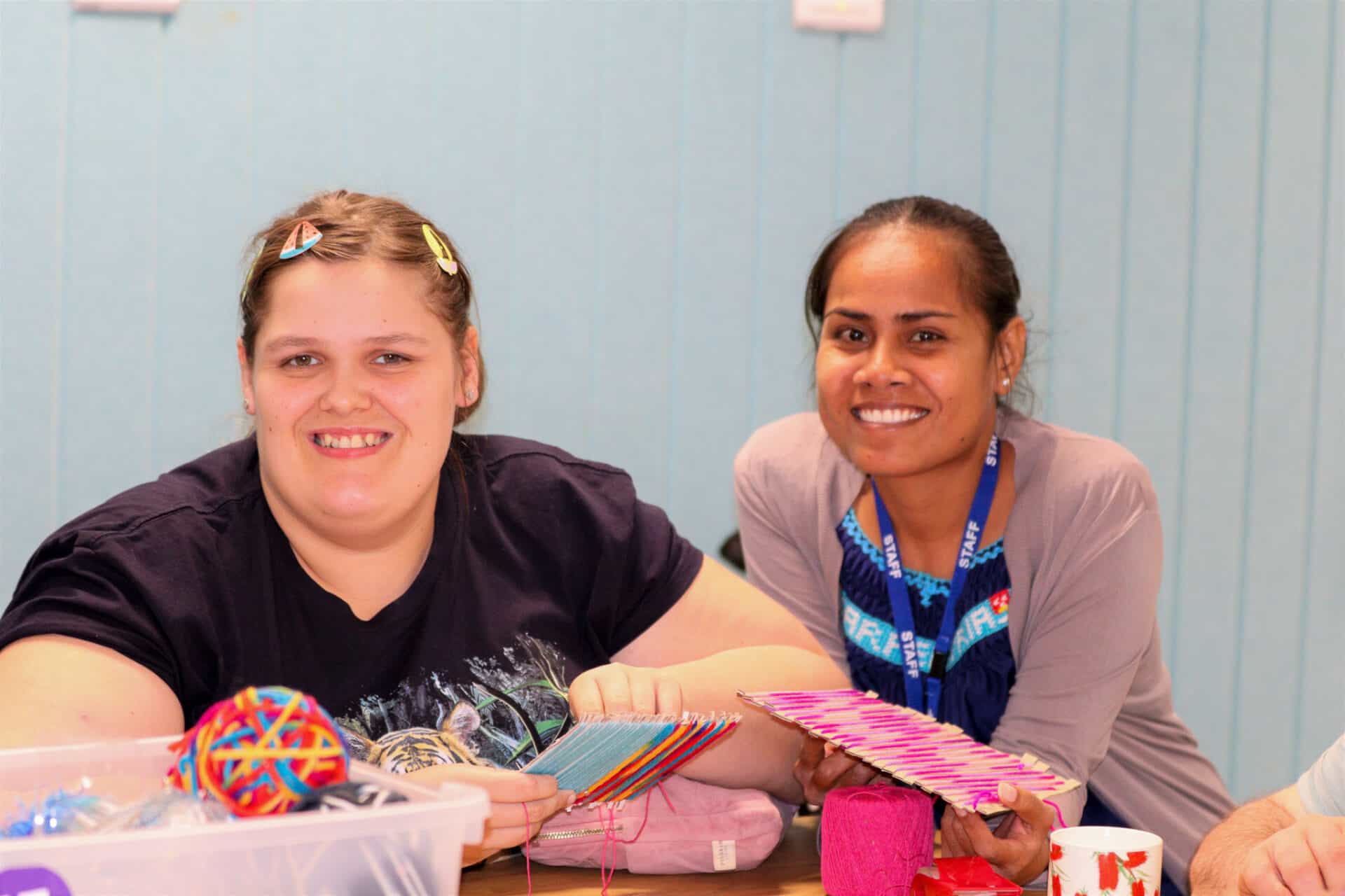 Two young women smile at the camera while holding a woollen craft activity.