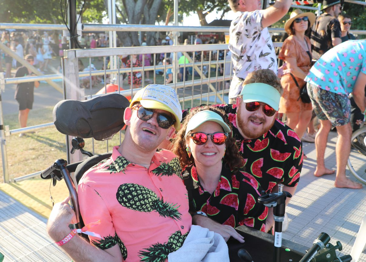 3 people in colourful shirts pose for a photo on the accessible viewing platform at Bass in the Grass. They are all wearing hats and sunglasses.