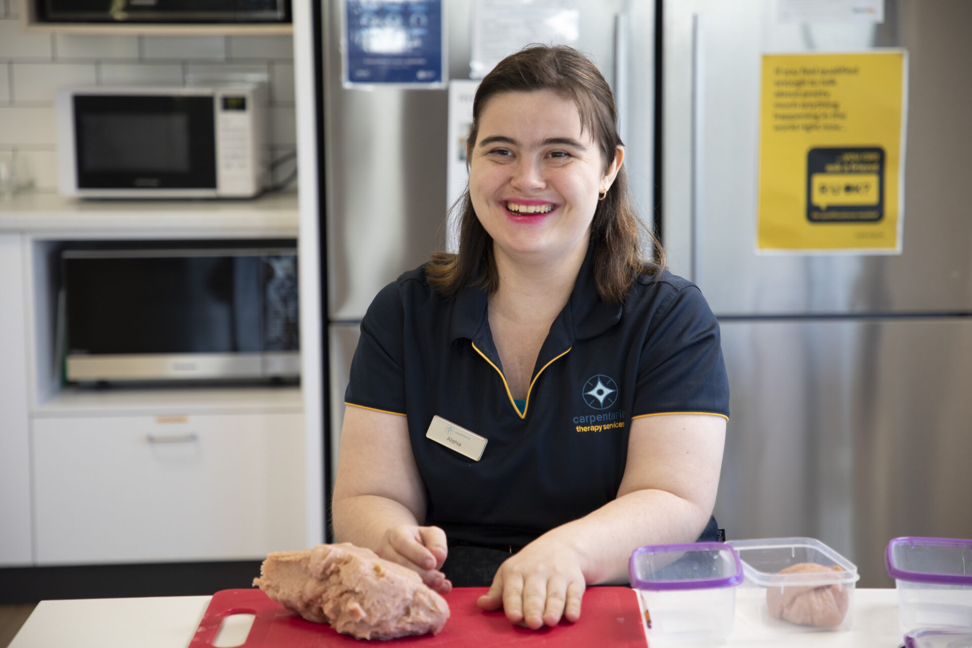 Alishia sits in a kitchen, making pink playdoh. She wears a Therapy Services polo shirt and is smiling