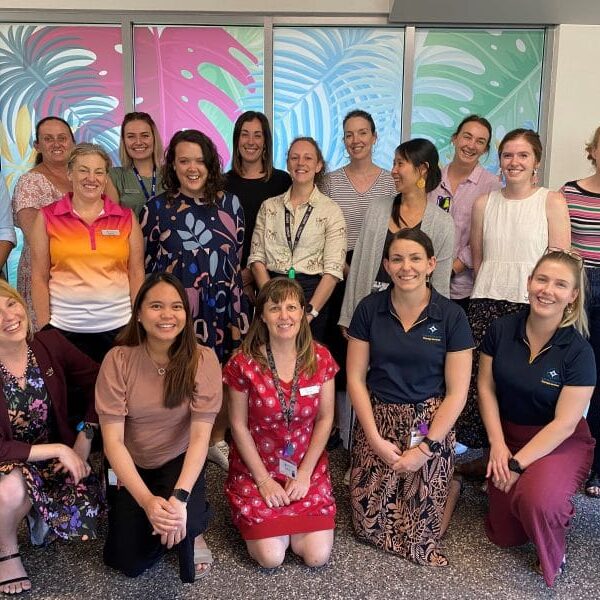A group of Allied Health professionals standing in front of a colourful background. They are smiling and laughing.