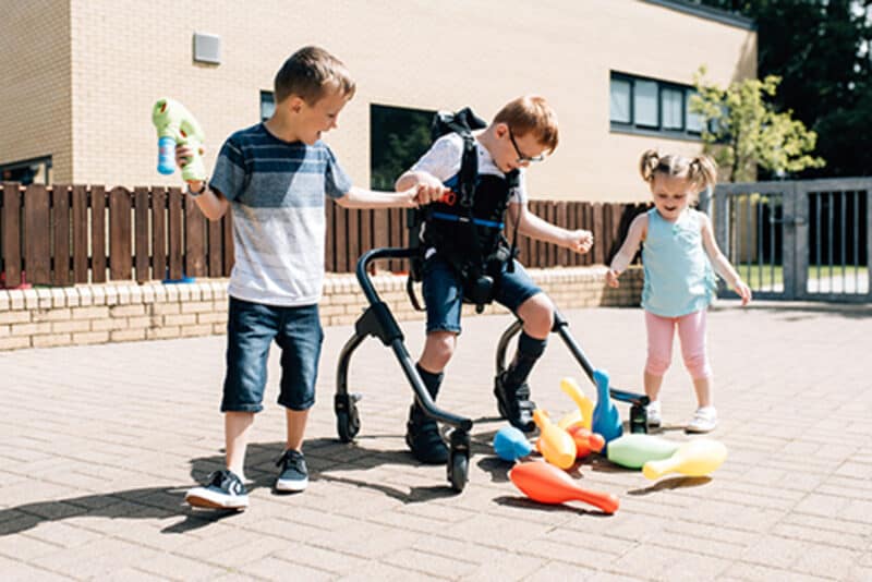 A child using a piece of standing equipment plays outdoors with 2 able-bodied children.